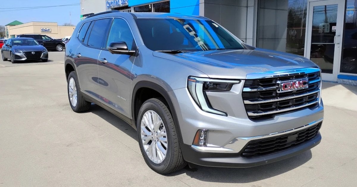 Silver 2026 GMC Acadia SUV parked at Jay's Chevrolet GMC's dealership lot in a three-quarter front view, with a showroom building, other vehicles, and a clear blue sky in the background.