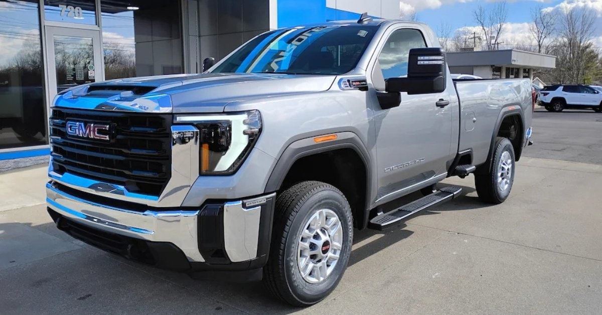  A silver 2026 GMC Sierra 2500 HD SLE Regular Cab pickup truck with a long bed, parked on a concrete lot in front of a modern dealership, Don's Chevrolet GMC. The truck features a black grille with a red GMC logo, C-shaped LED headlights, chrome and black accents, and large towing mirrors. The background includes a dealership with glass windows, blue accents, and other vehicles, set against a bright blue sky with scattered clouds.