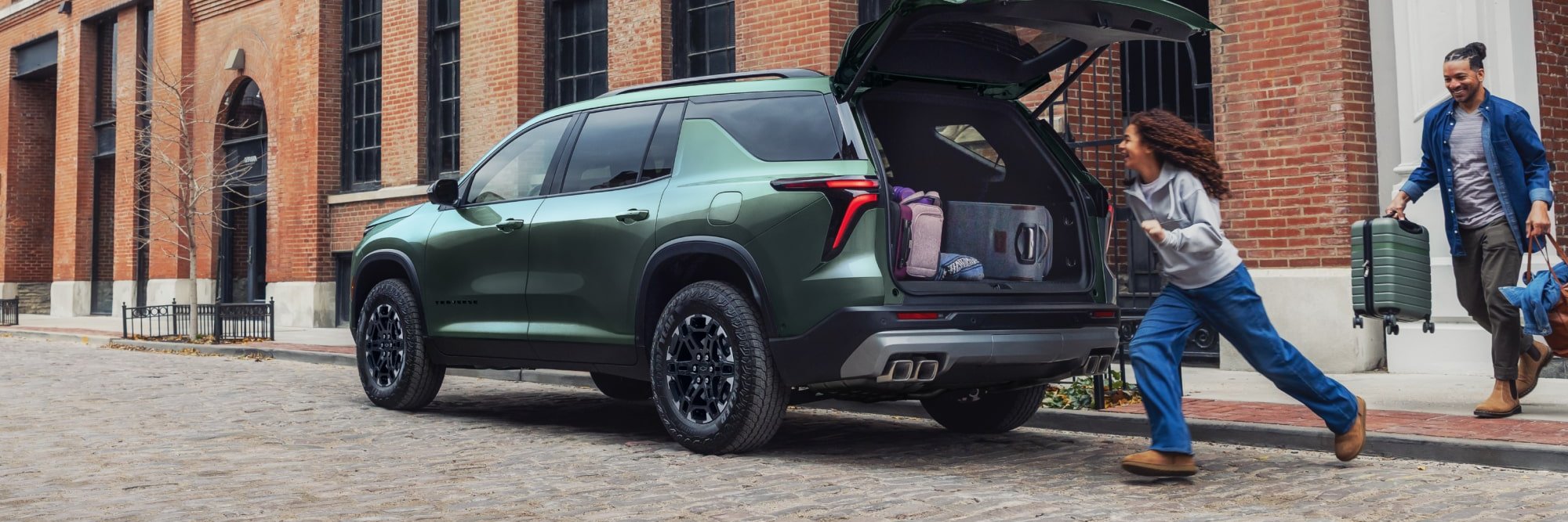 A green Traverse Z71 parked on a brick street with its liftgate open as a girl runs to load her bags