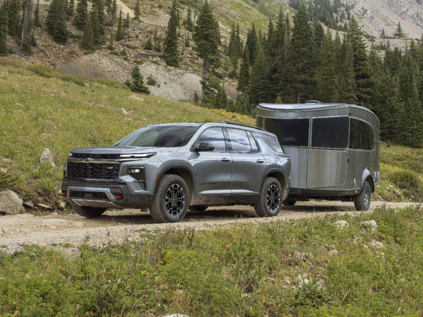 A gray Traverse towing a small travel trailer on a mountain dirt road lined with pines
