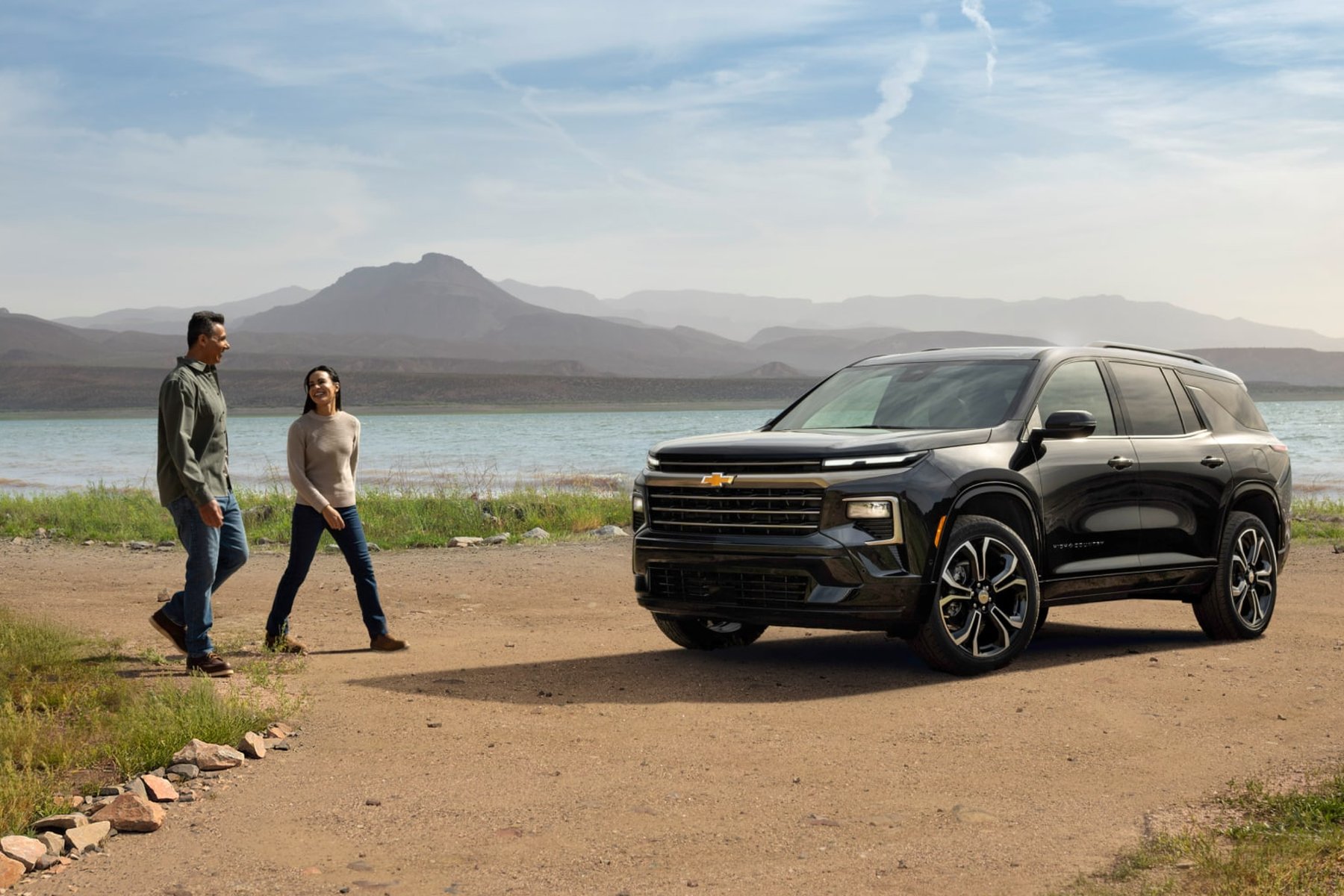 A couple walking toward a black Chevrolet Traverse parked on a dirt lot beside a mountain lake