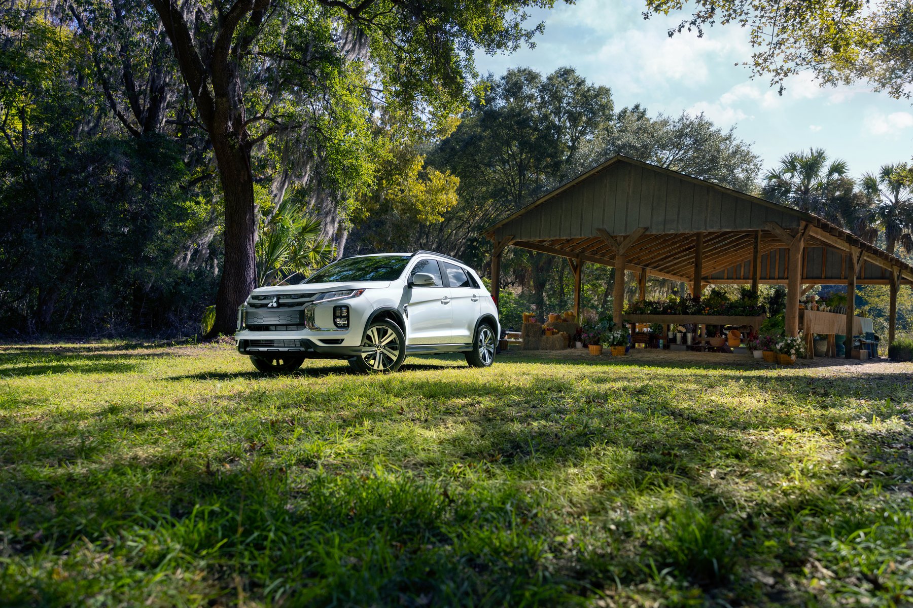 A white Mitsubishi Outlander Sport parked on a lawn in front of a wooden market pavilion, under large trees