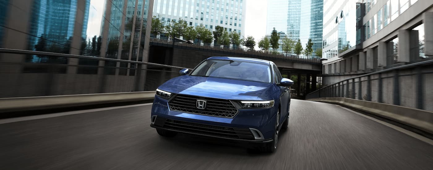 A blue 2023 Honda Accord Sport Hybrid is shown from the front driving on a city street.