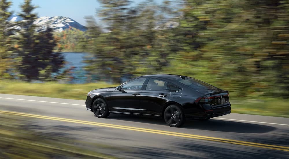 A black 2023 Honda Accord Sport is shown from the side driving on a tree-lined road.