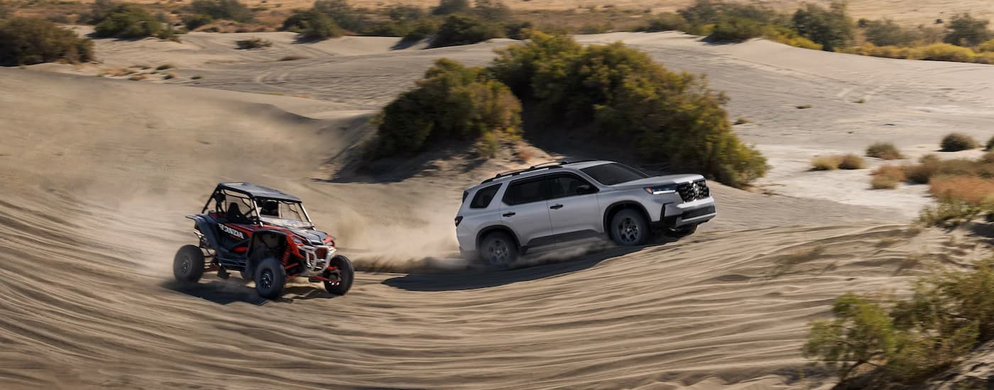 A silver 2023 Honda Pilot Trailsport is shown from the side off-roading on sand next to a UTV.