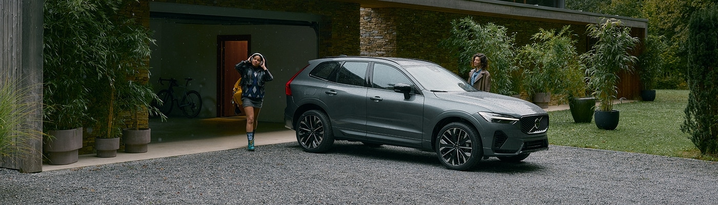 A woman and girl approaching a 2026 Volvo XC60 Hybrid parked outside a modern rustic home