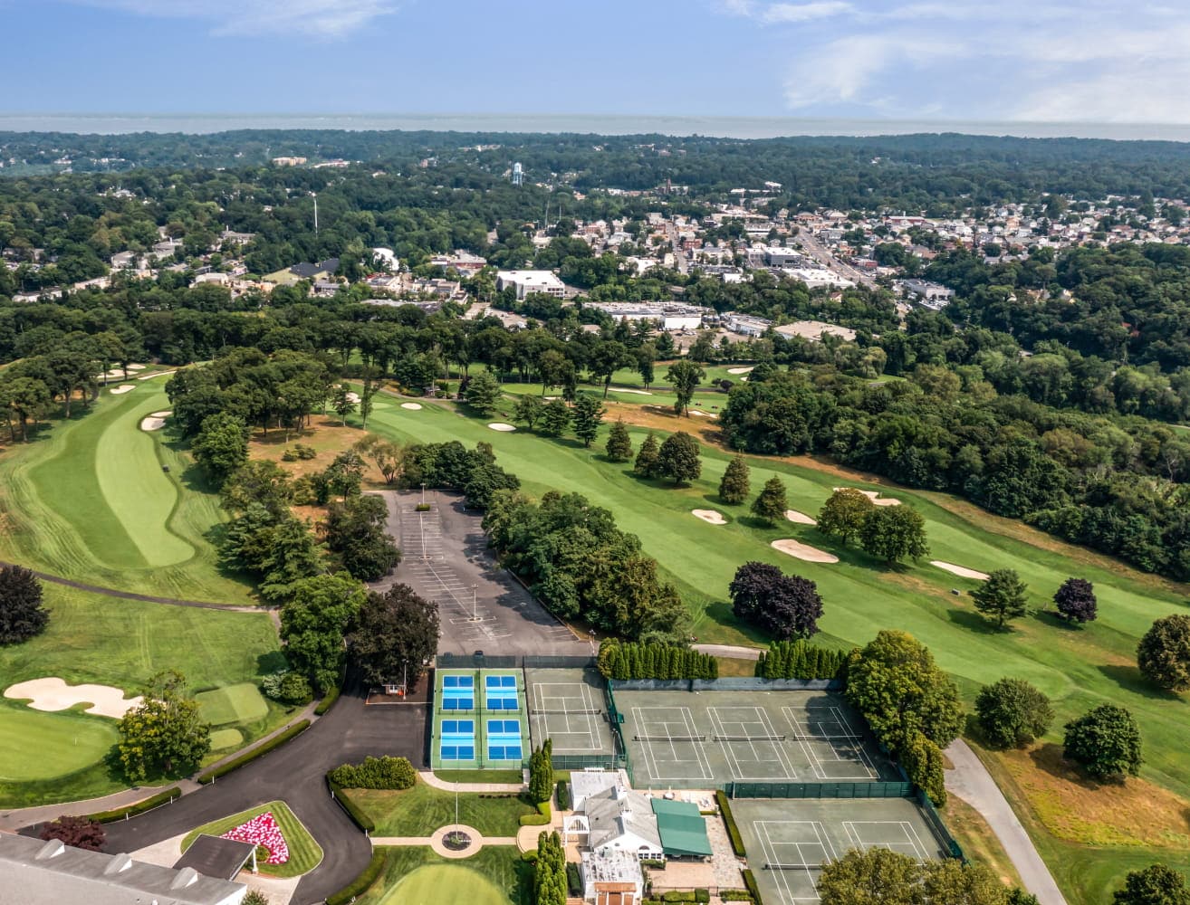 Aerial view of a tree-lined community featuring a golf course, tennis courts, and surrounding neighborhoods.