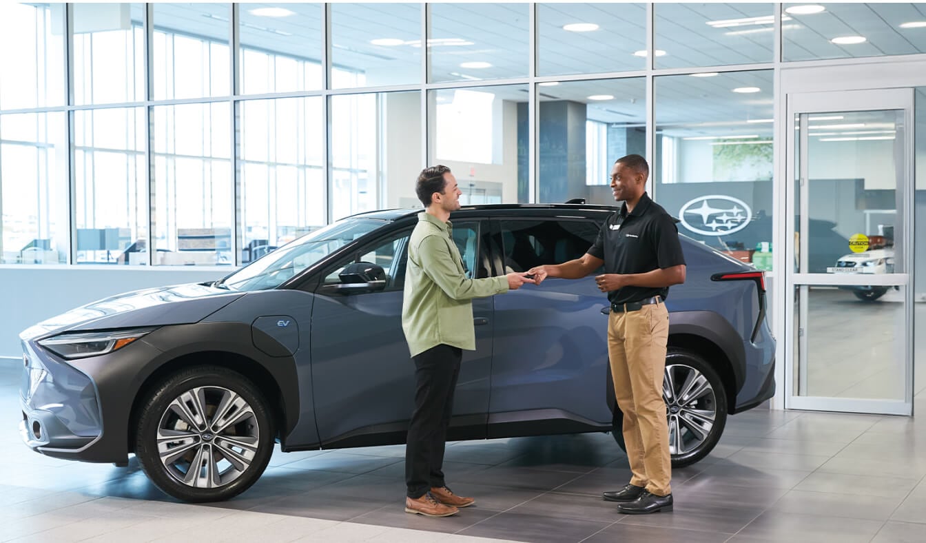 A sales representative handing over vehicle keys to a customer inside a modern Subaru showroom, standing beside a 2026 Subaru Solterra.