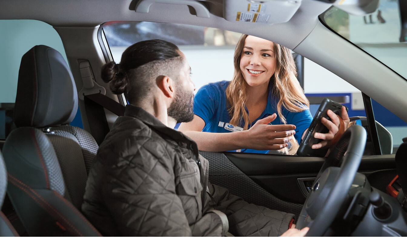 A Subaru service advisor holding a digital tablet and speaking with a male driver seated inside a 2026 Subaru Outback in a service check-in lane.