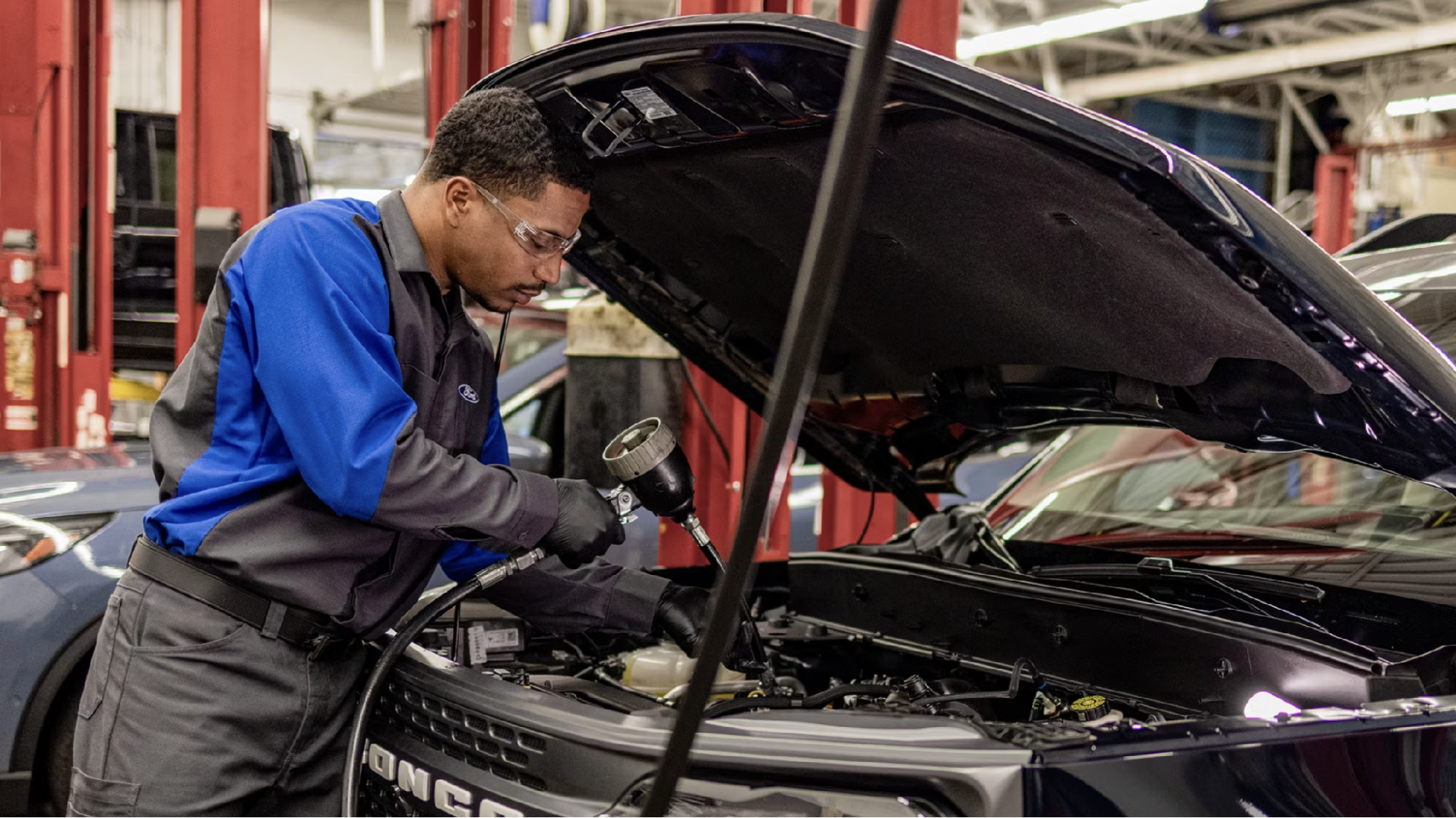 Ford technician in blue and gray uniform working under the open hood of a Ford Bronco in a service bay.