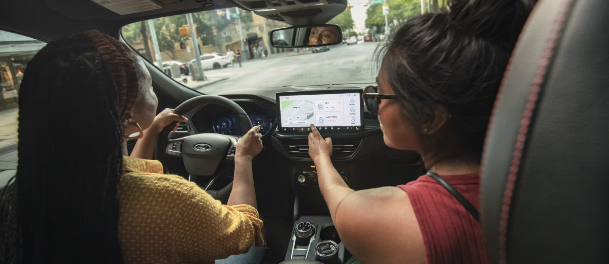Two friends inside a Ford Escape using the touchscreen infotainment system while driving through the city