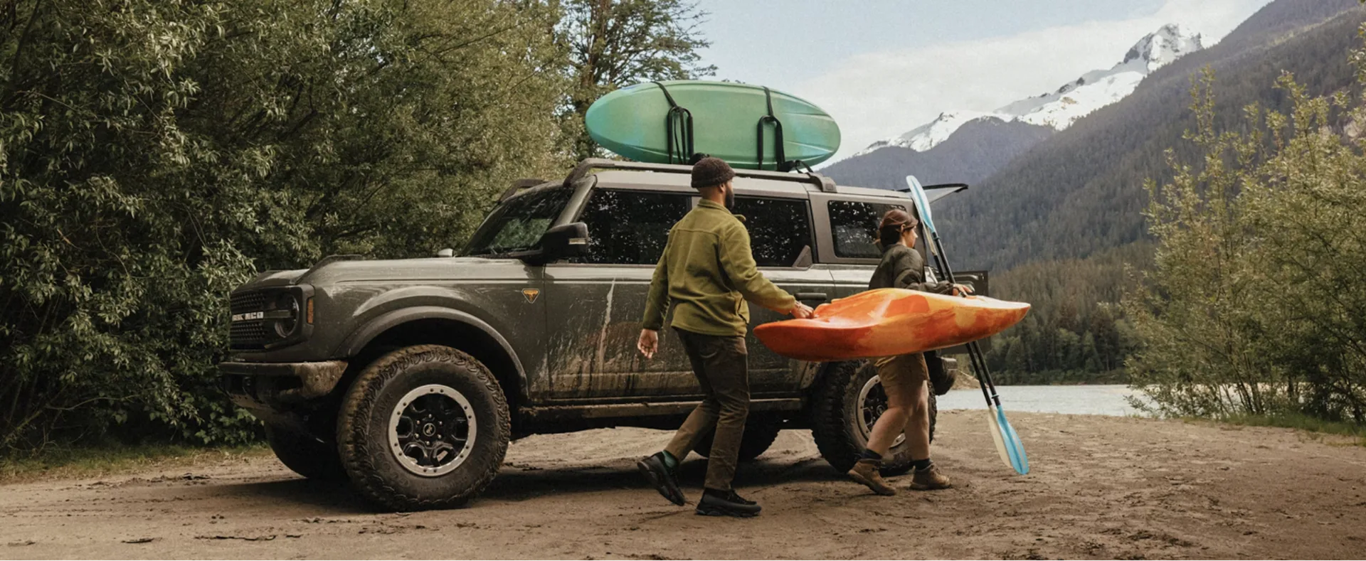 Two people loading kayaks onto a mud-covered Ford Bronco with a roof rack near a river and a mountain