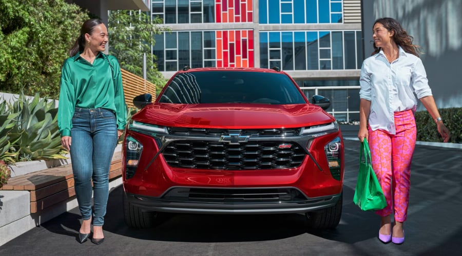 Red 2026 Chevy Trax RS subcompact crossover SUV parked outside a modern shopping plaza in Marlton, NJ, with two local commuters walking alongside it.