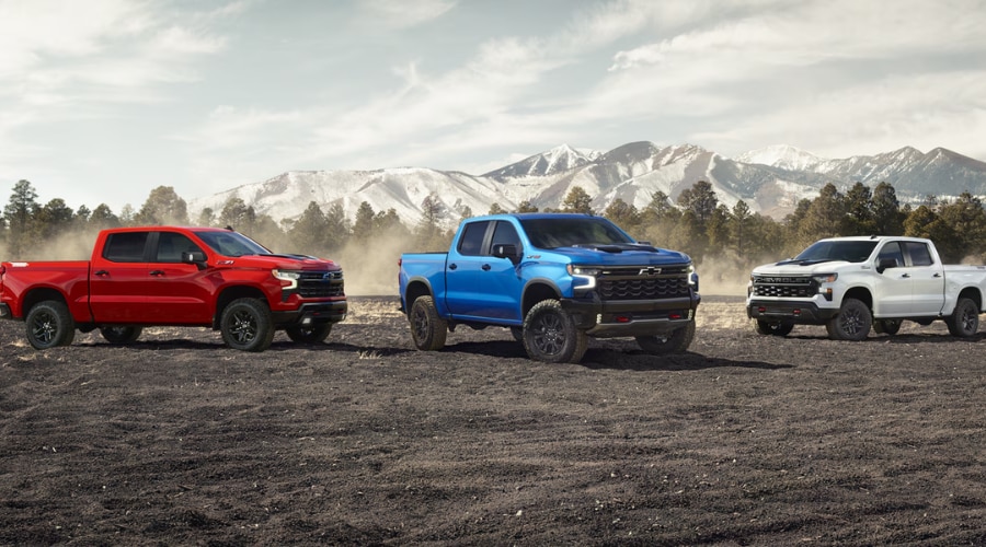 Lineup of three CarBravo certified Silverado 1500 trucks in red, electric blue, and summit white parked on dirt terrain with mountain backdrop, showcasing pre-owned truck inventory for South Jersey drivers in Marlton and Deptford, NJ.