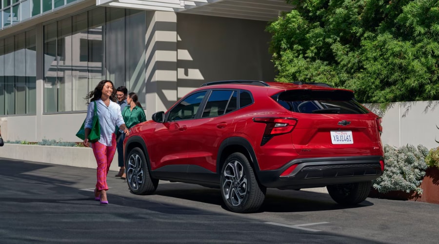 Rear three-quarter view of a bright red 2026 Chevy Trax RS parked at a suburban shopping center, showcasing the rear bumper parking sonar sensors and Chevy Safety Assist suite available at Elkins Chevrolet in Marlton, NJ.