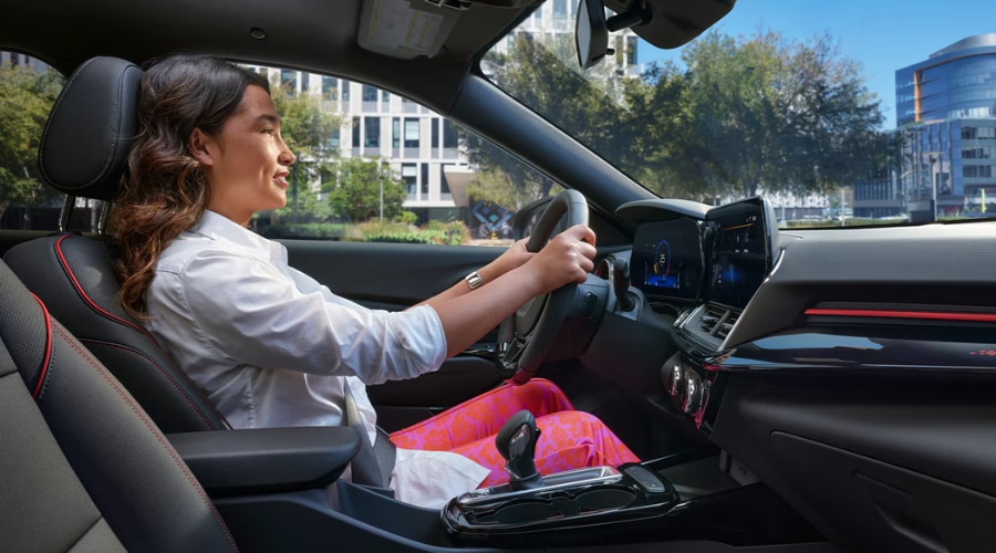 Side profile view of a woman driving a 2026 Chevy Trax RS, showing the ergonomic seating position, leatherette seats with red stitching, and clear line-of-sight to the dual cabin displays at Elkins Chevrolet in Marlton, NJ.