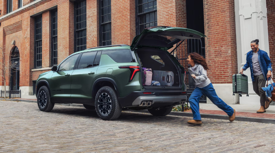 Rear profile of a green 2027 Chevrolet Traverse Z71 with its open AutoSense Power Liftgate as a family loads luggage into the spacious 98 cubic foot cargo area, available at Elkins Chevrolet in Marlton, NJ.