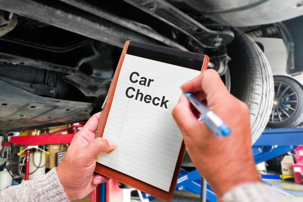 A person holding a clipboard with Car Check written on it, standing under a raised vehicle in a car repair workshop