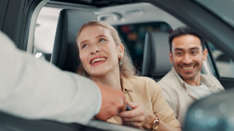 A car dealer handing a car key to two customers sitting inside a vehicle
