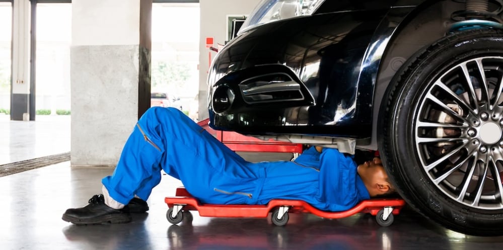 A person with light brown skin in a blue jumpsuit lying underneath the front of a vehicle 