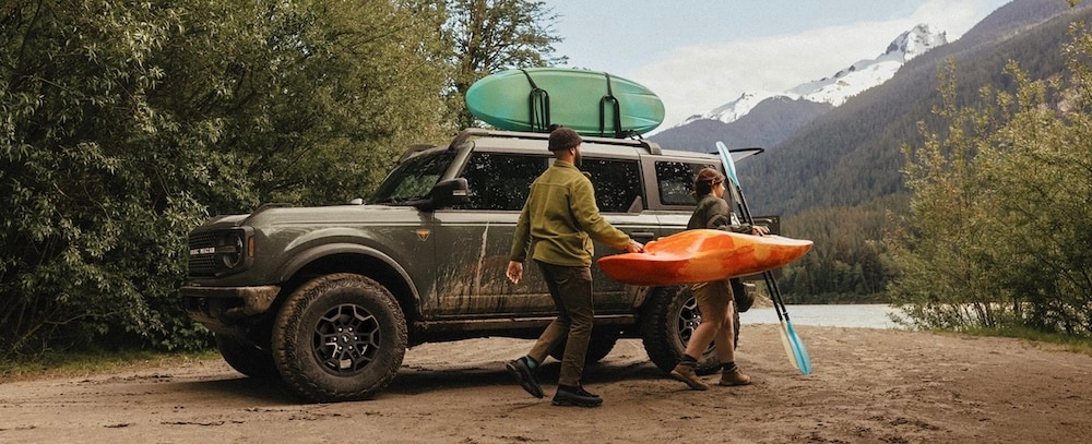 Two people carrying a kayak beside a 2026 Ford Bronco parked beside a river
