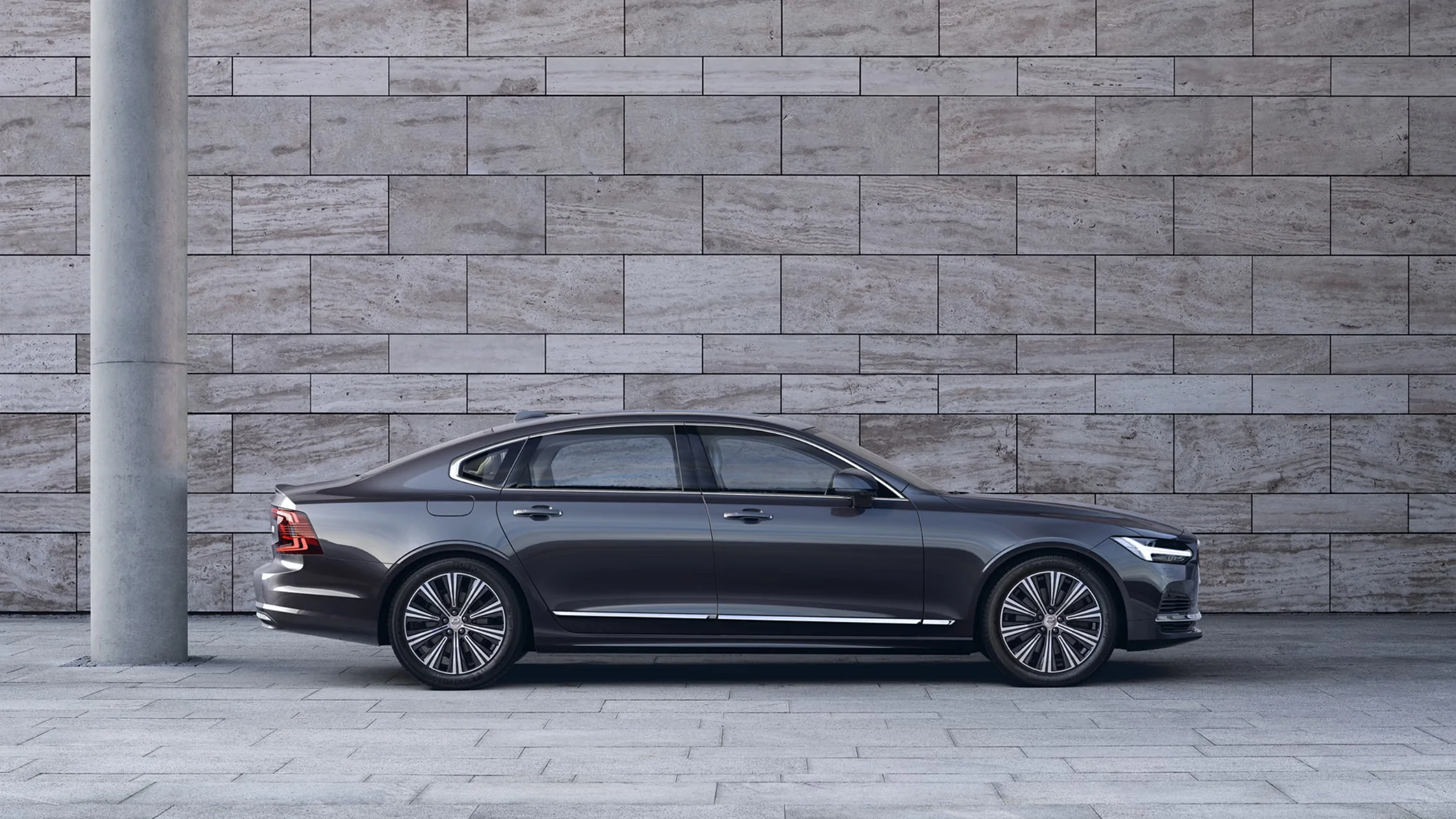 Side view of a gray 2025 Volvo S90 parked in front of a tiled concrete wall with modern urban styling.