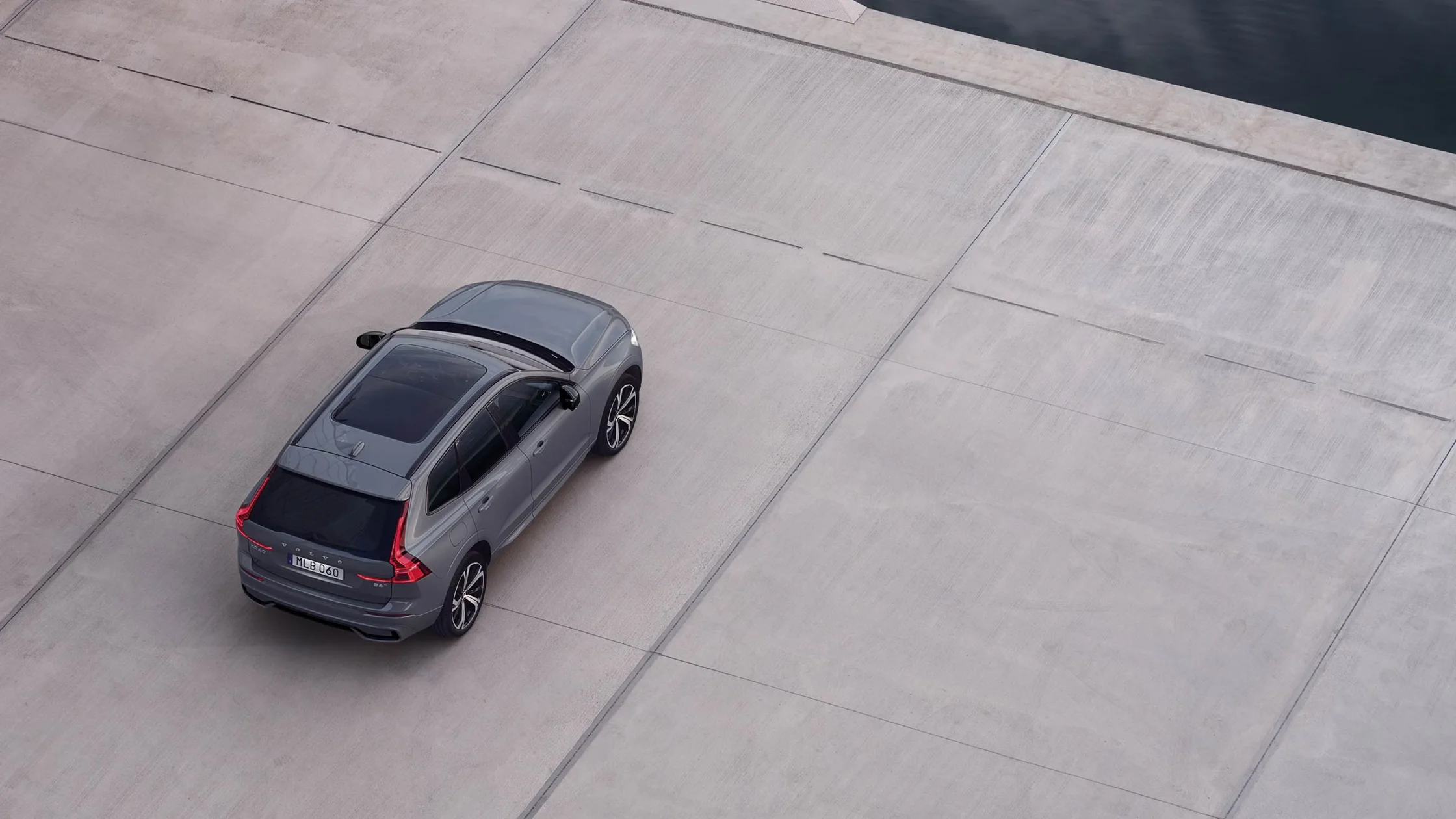Gray 2025 Volvo XC60 SUV parked on a large, clean concrete surface near a waterfront area.