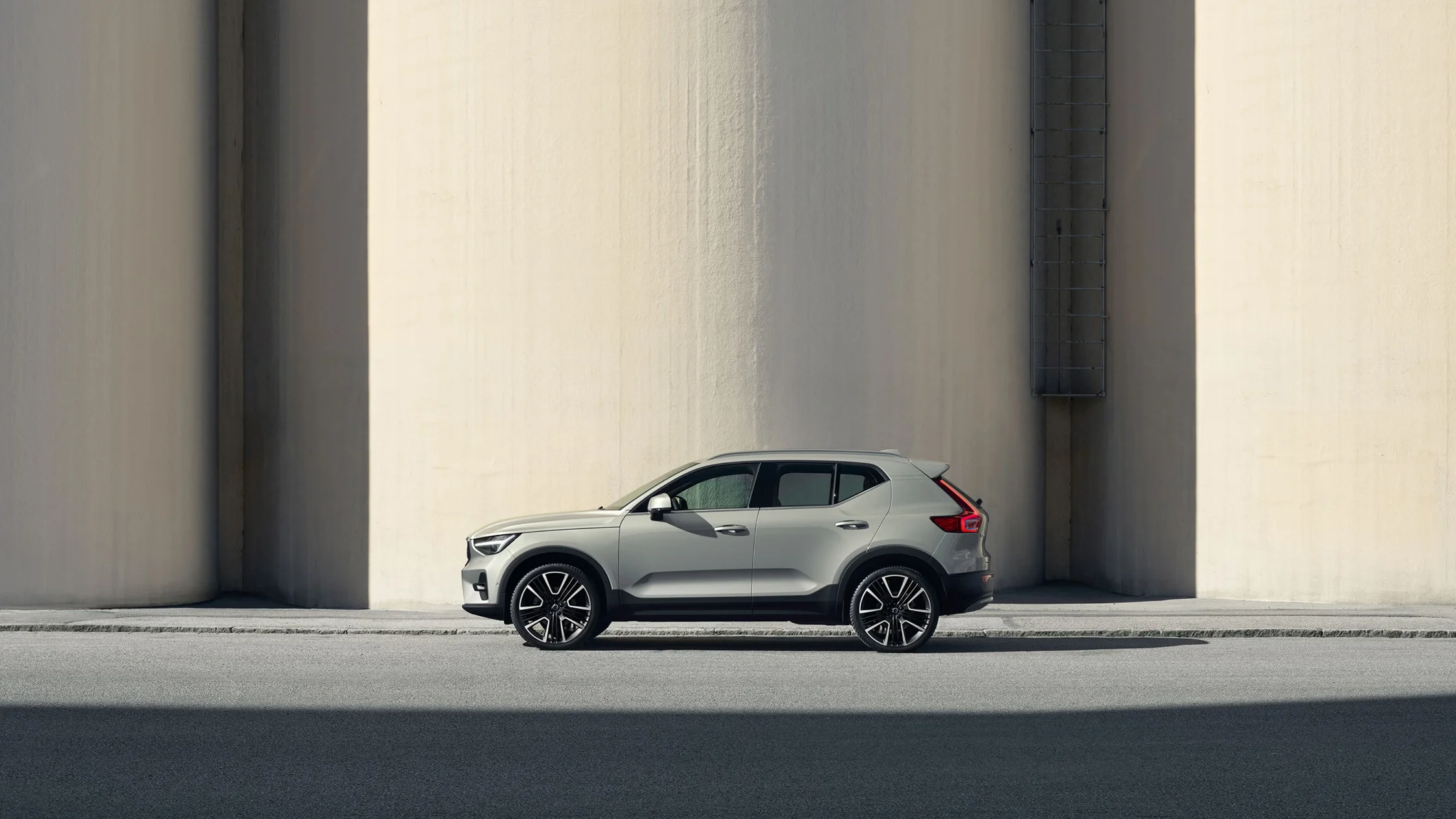 Profile of a light gray 2025 Volvo XC40 SUV parked along a city street with a curved concrete backdrop.