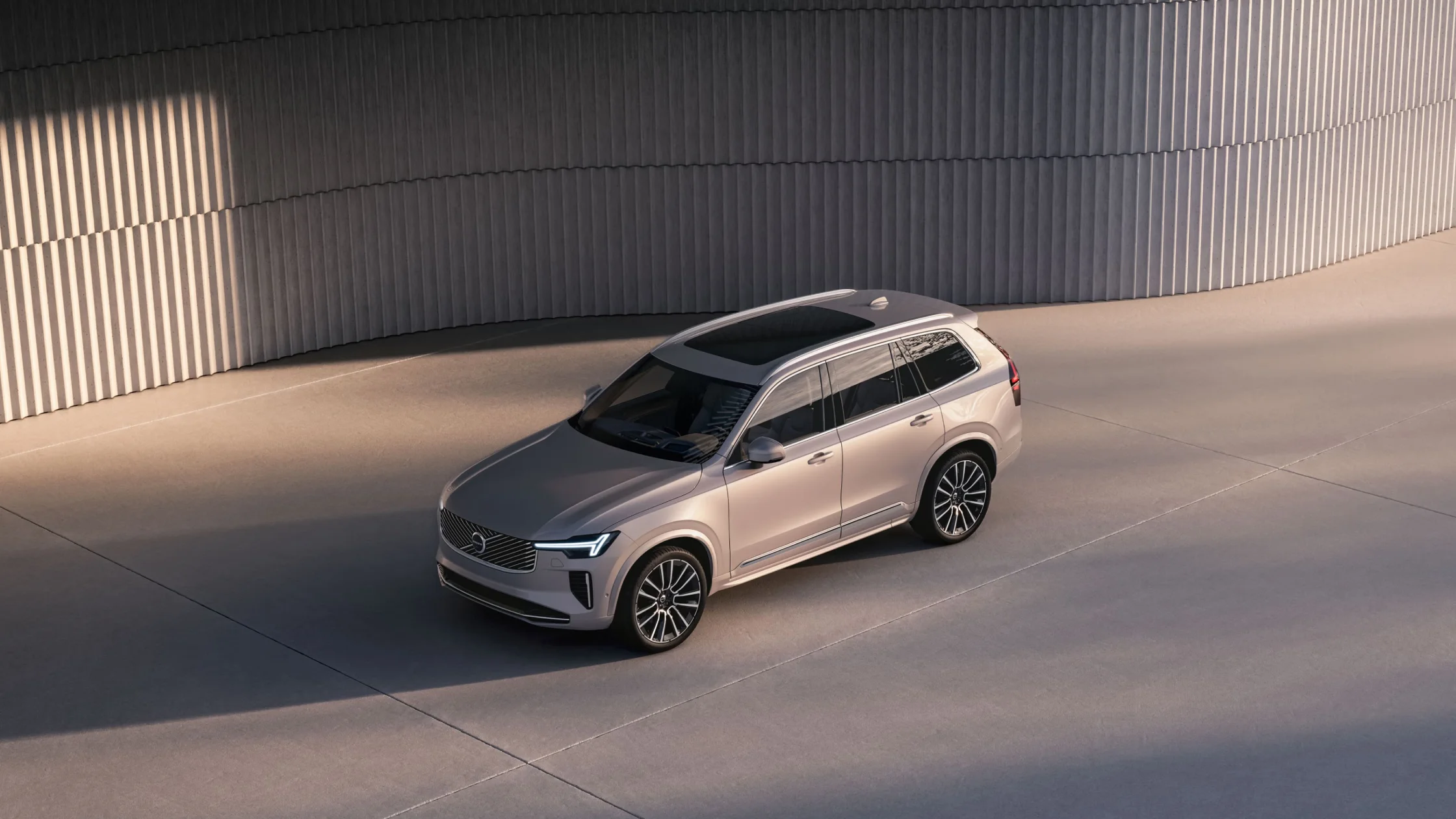 Top-side view of a silver 2025 Volvo XC90 with panoramic roof parked in modern architectural surroundings.
