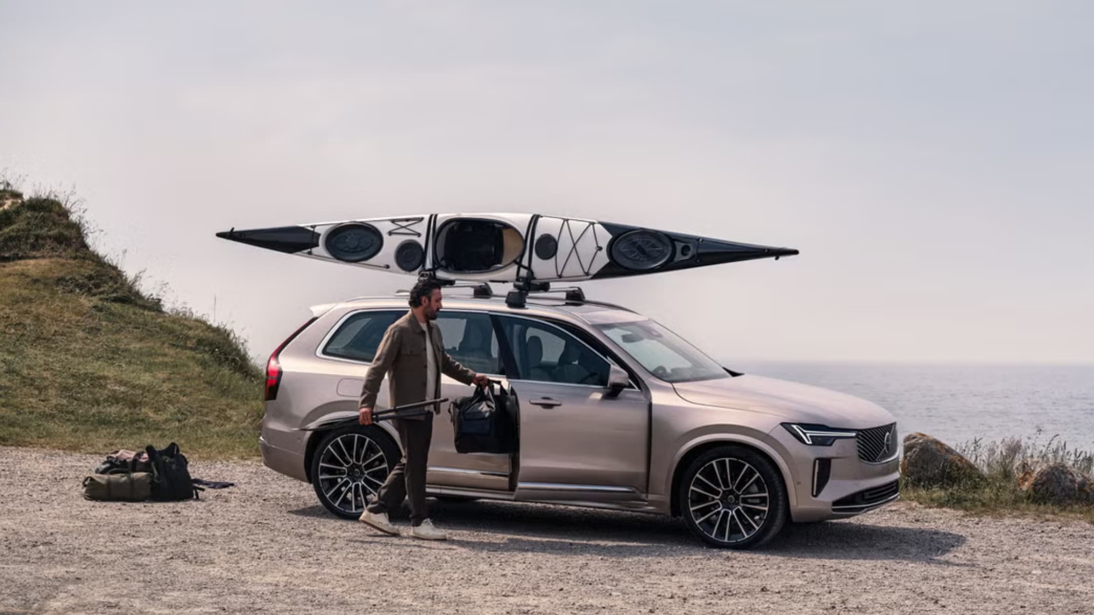 Beige 2025 Volvo XC90 Plug-In Hybrid with a kayak on the roof, parked by a cliffside with the driver unloading gear.