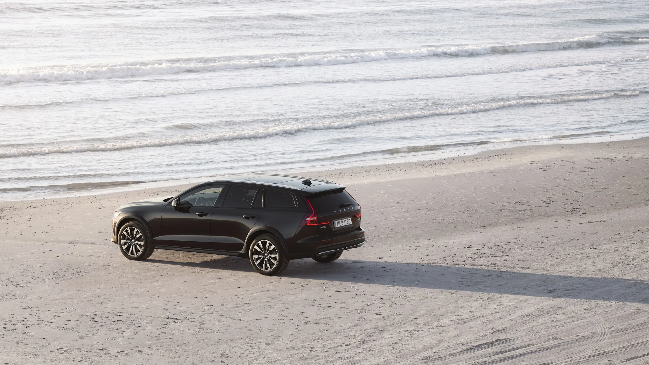 Black 2025 Volvo V60 Cross Country wagon parked on a beach near the ocean with waves in view.