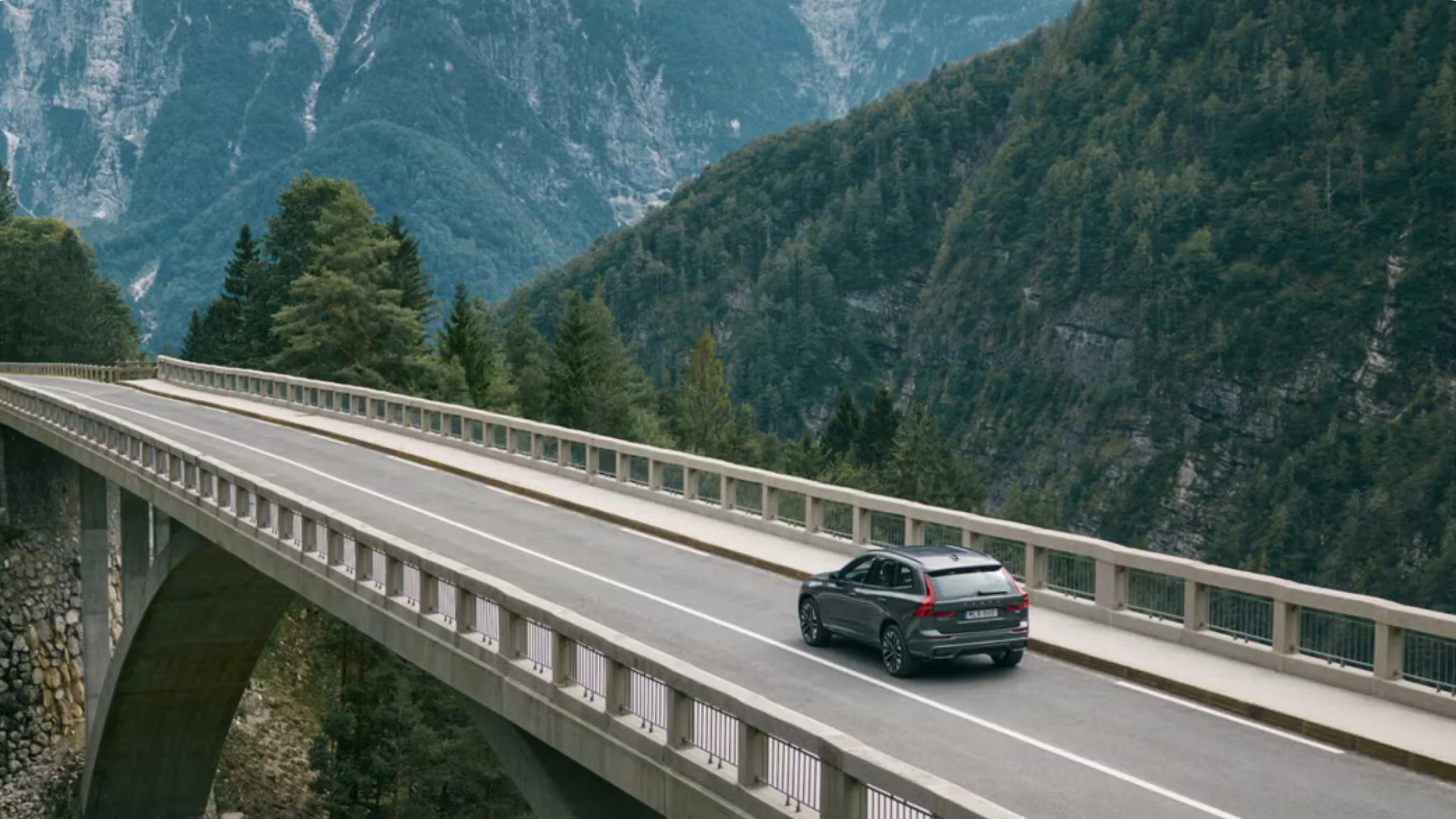 2025 Volvo XC60 driving across a mountain bridge with scenic forest cliffs in the background