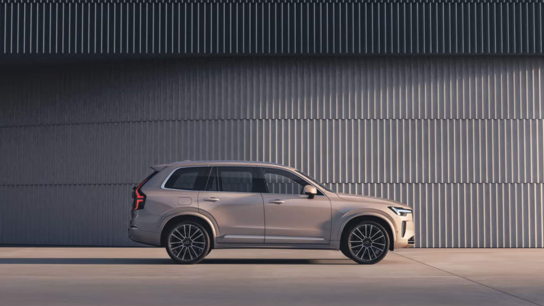 Side profile of a beige 2025 Volvo XC90 SUV parked in front of a modern metal wall under natural daylight.