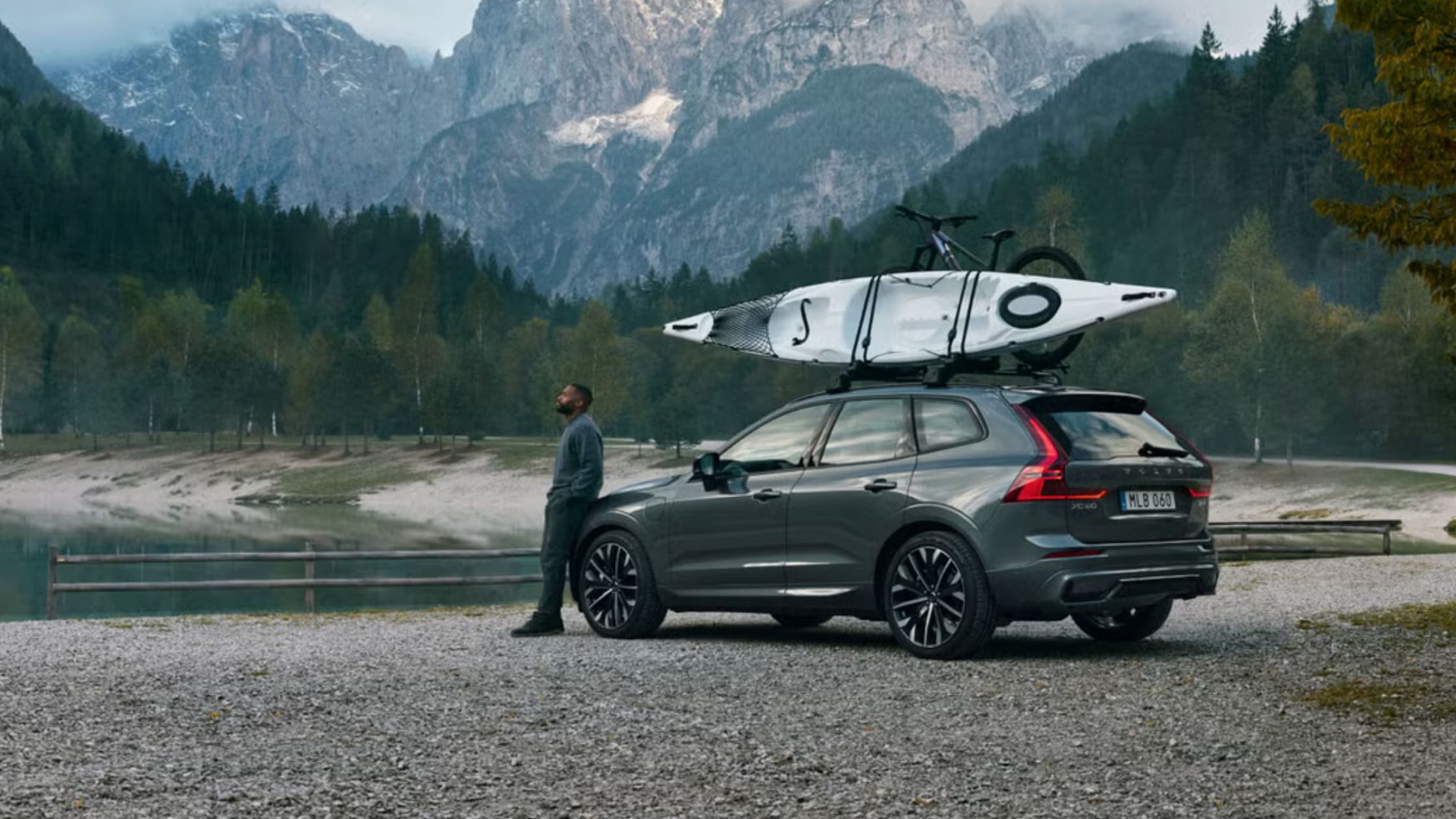 Gray 2025 Volvo XC60 Plug-In Hybrid parked by a lake with a kayak and bicycle mounted on the roof rack.