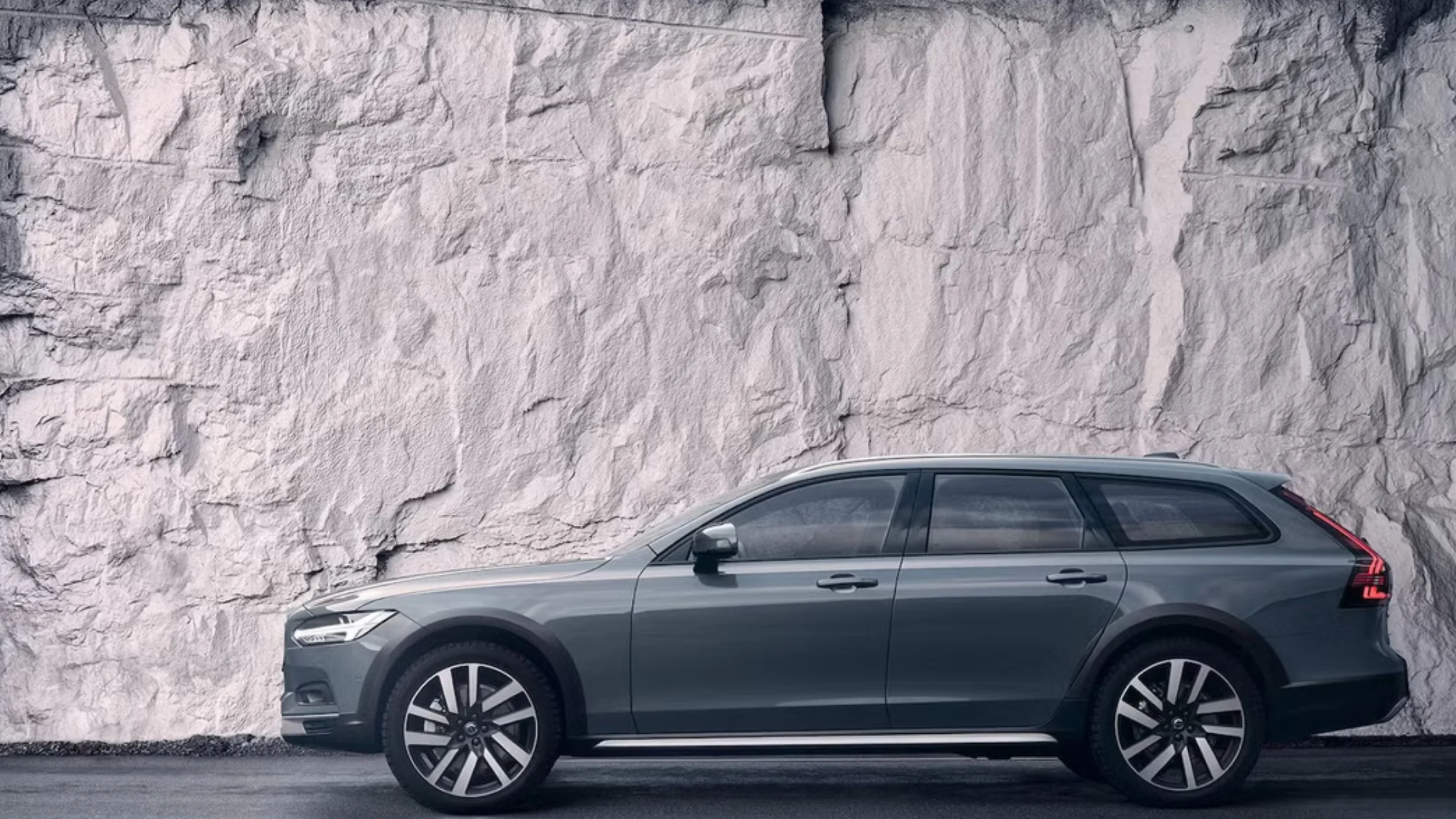 Side view of a 2025 Volvo V90 Cross Country wagon in gray parked beside a rock wall.
