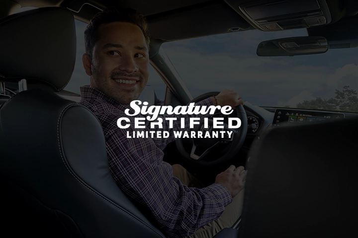 Signature Certified Limited Warranty logo overlayed on an image of a smiling man in the driver seat of a Honda looking over his shoulder through the back window