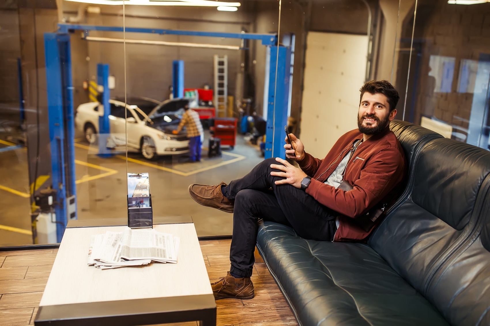 A customer sitting on the couch in a service lounge with his phone. A service member works on a vehicle in the background.