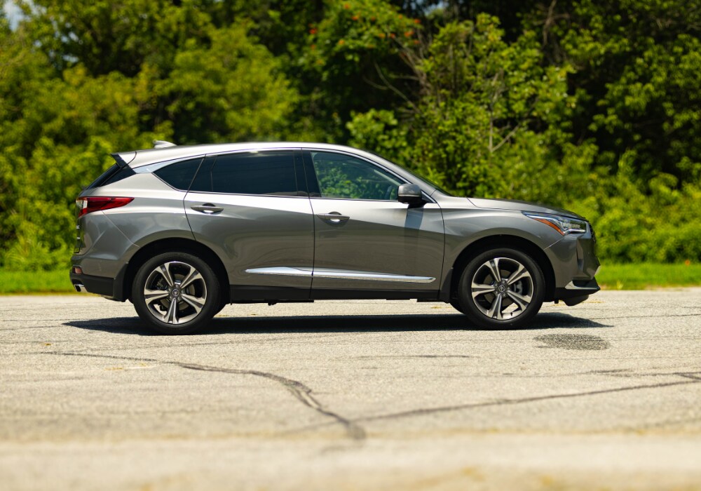 A side view of a new Acura RDX. The RDX is silver and parked in a parking lot with landscaping in the background
