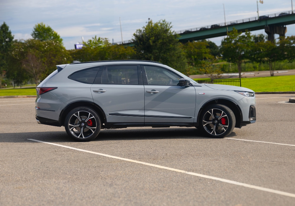 Gray 2025 Acura MDX parked on a paved surface with a bridge in the background