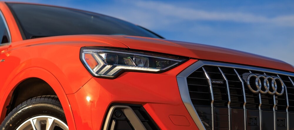 Close up photo of the front of a 2024 Audi Q3 SUV in the color orange. The Audi rings are visible on the front logo