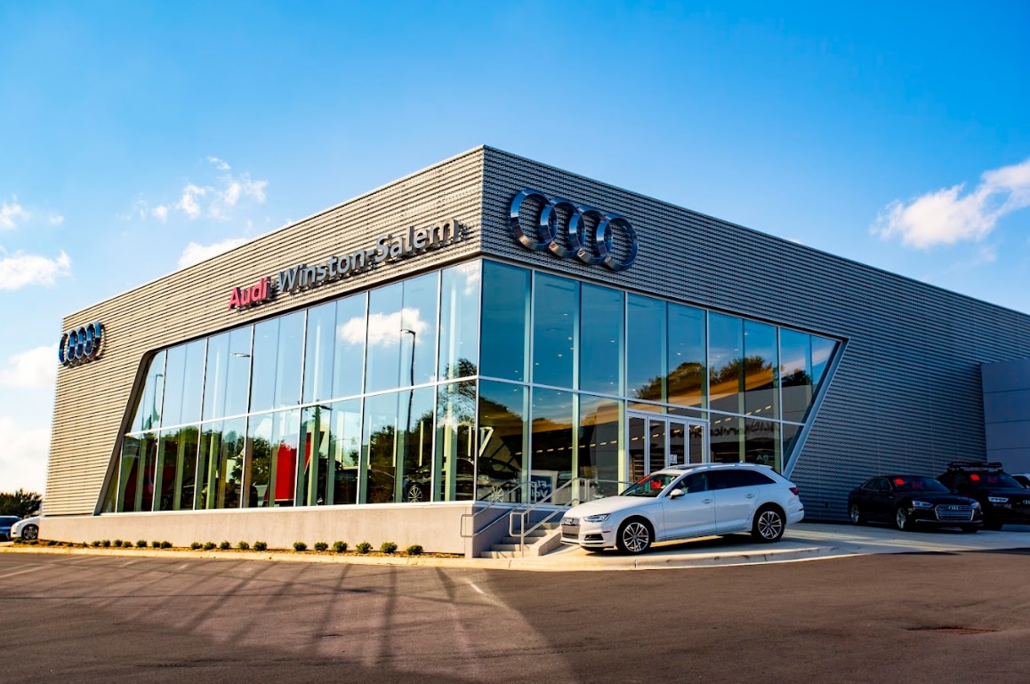 exterior photo of the Audi Winston Salem dealership in Winston Salem NC. There is a white Audi vehicle parked outside the dealership