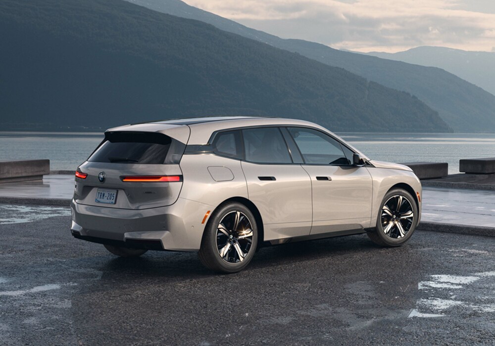 bmw-ix-performance.png Rear exterior view of a 2025 BMW iX parked on a paved surface with mountains and a large body of water in the background
