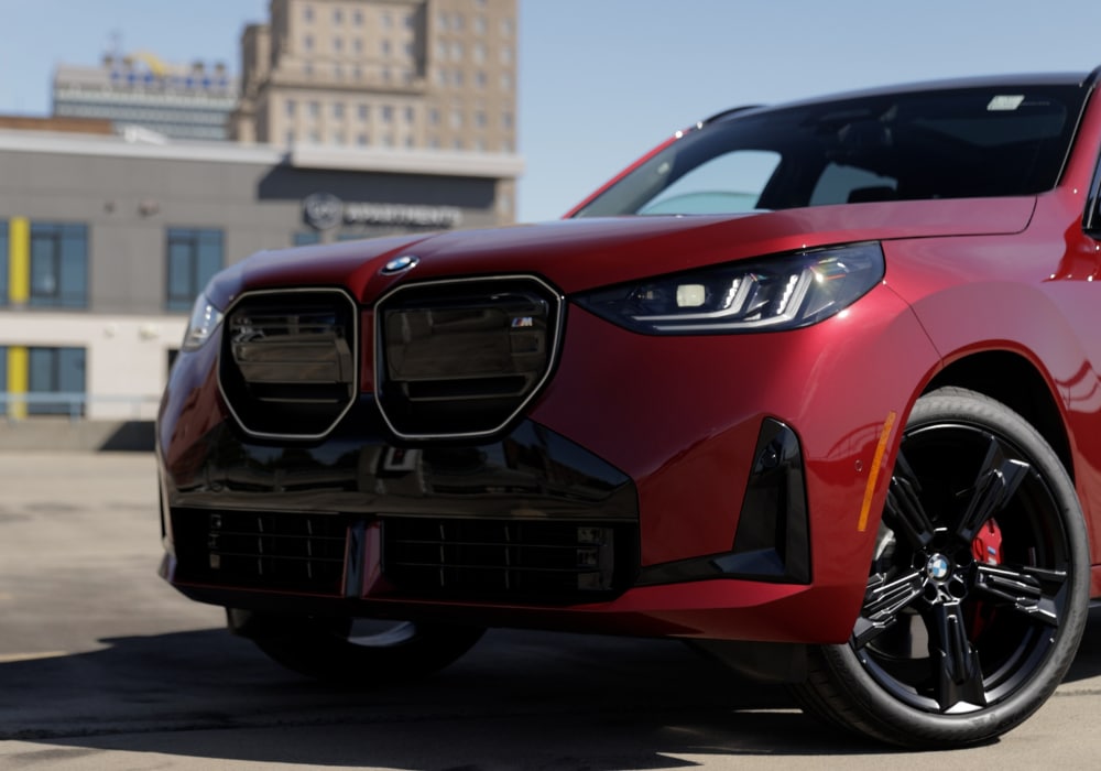 2025-bmw-x3-for-sale.png Front half of a red 2025 BMW X3 parked on the top floor of a parking deck. There are other tall buildings in the background