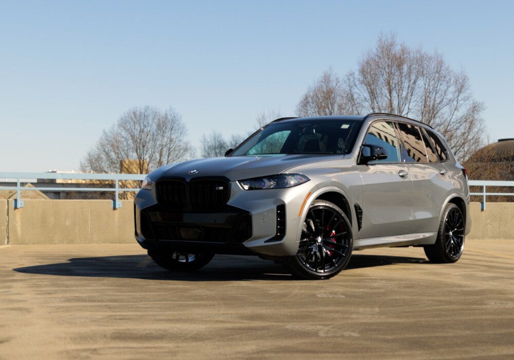 Gray 2025 BMW X5 SUV parked on a paved surface with a few trees in the background