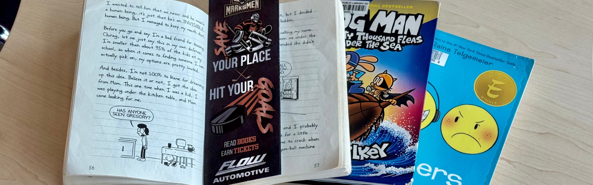Marksmen reading achievemen bookmark laying in the middle of an open book. There are several books in a pile