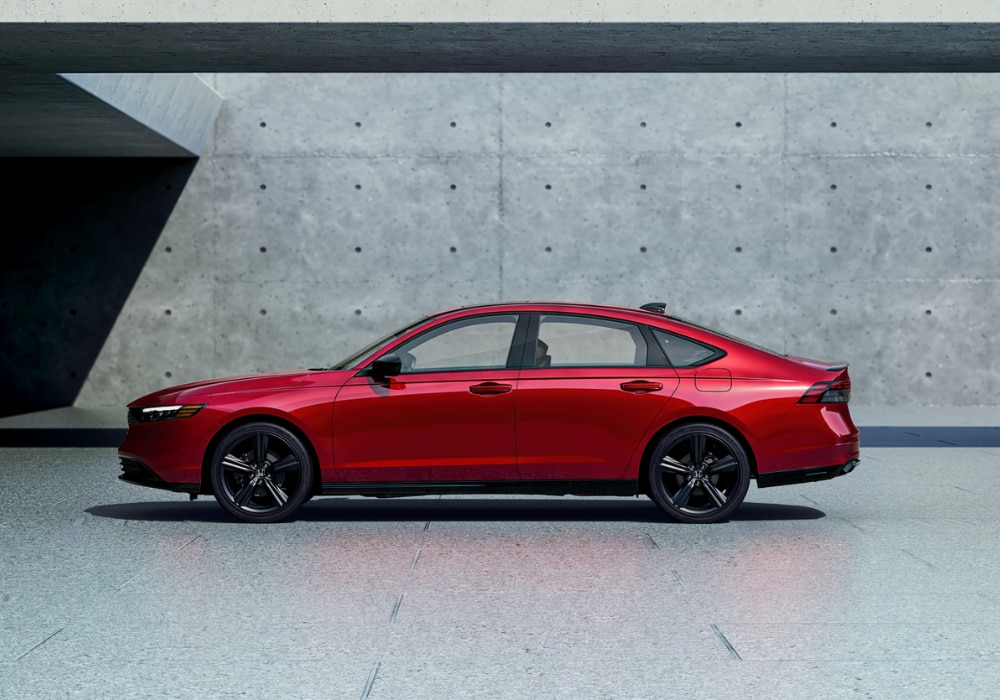 Driver's side view of a red Honda Accord Hybrid parked in front of an industrial building