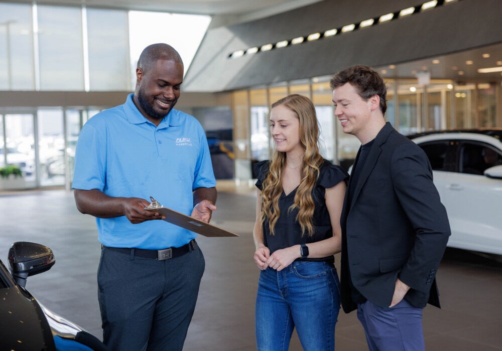 Two customers standing next to a Flow employee inside a dealership. The employee is showing a clipboard to the customers