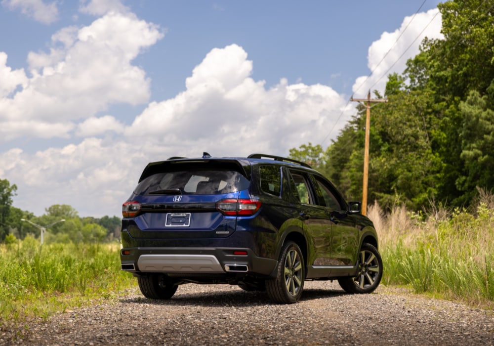 Rear exterior view of a new Honda Pilot parked on a gravel road. The SUV is blue and there are trees and clouds in the background