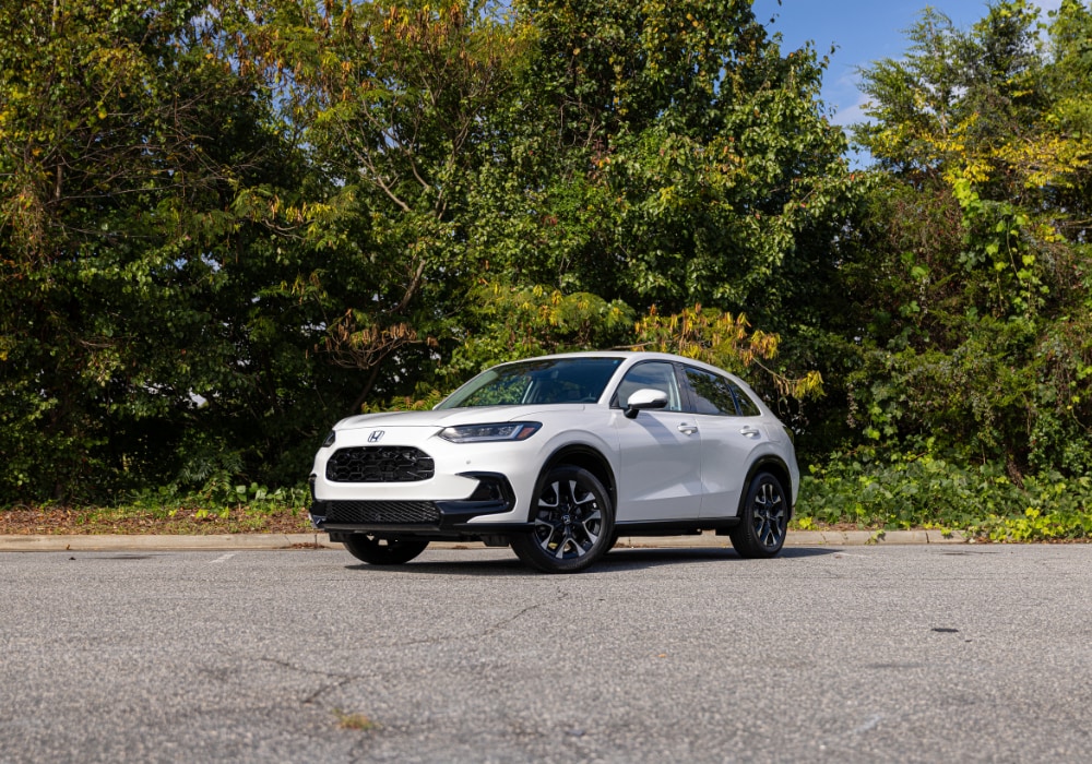 White 2025 Honda HR-V SUV parked on a paved surface with a wooded area in the background