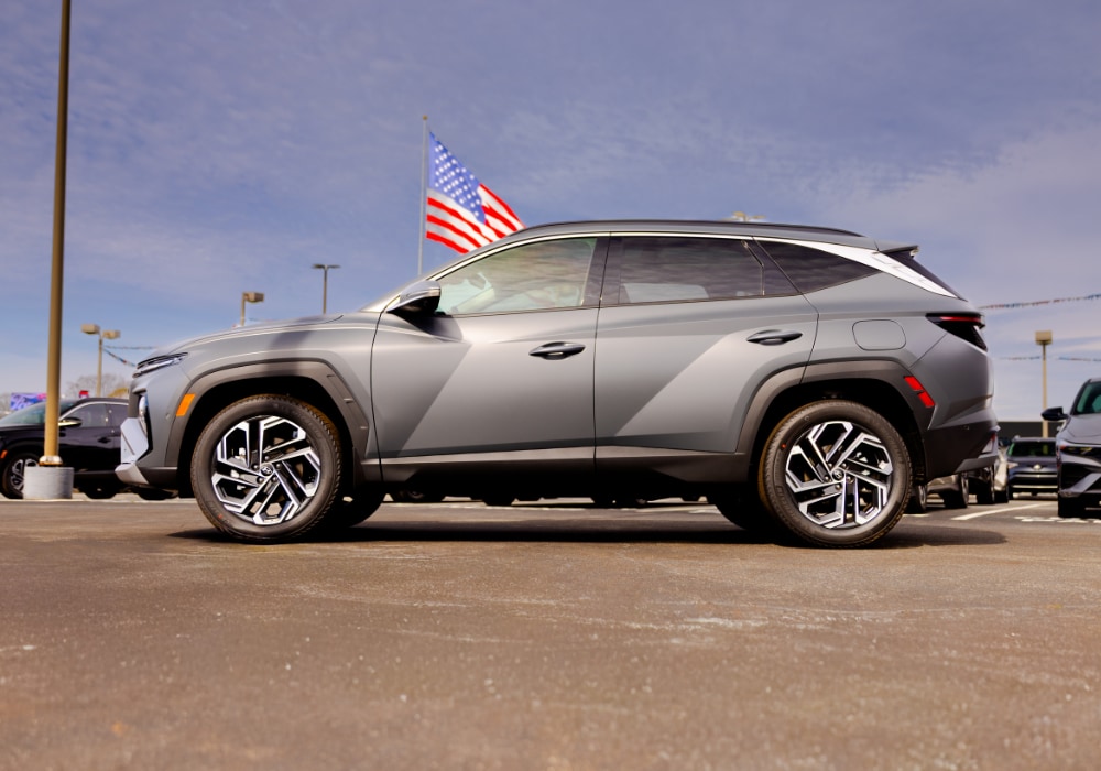 Driver's side exterior view of a 2026 Hyundai Tucson SUV. There is an American flag in the background
